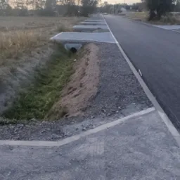 Nowa asfaltowa droga z wybrukowanym poboczem i rowem melioracyjnym z betonowymi wlotami. Widok na wprost, w tle zabudowania i drzewa.