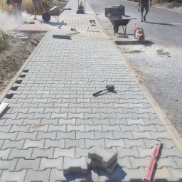 RATBUD - Układanie szarej kostki brukowej na chodniku w Szydłowcu. W tle pracownicy i narzędzia: taczka, zagęszczarka, piła do cięcia kostki. Widoczny postęp prac.