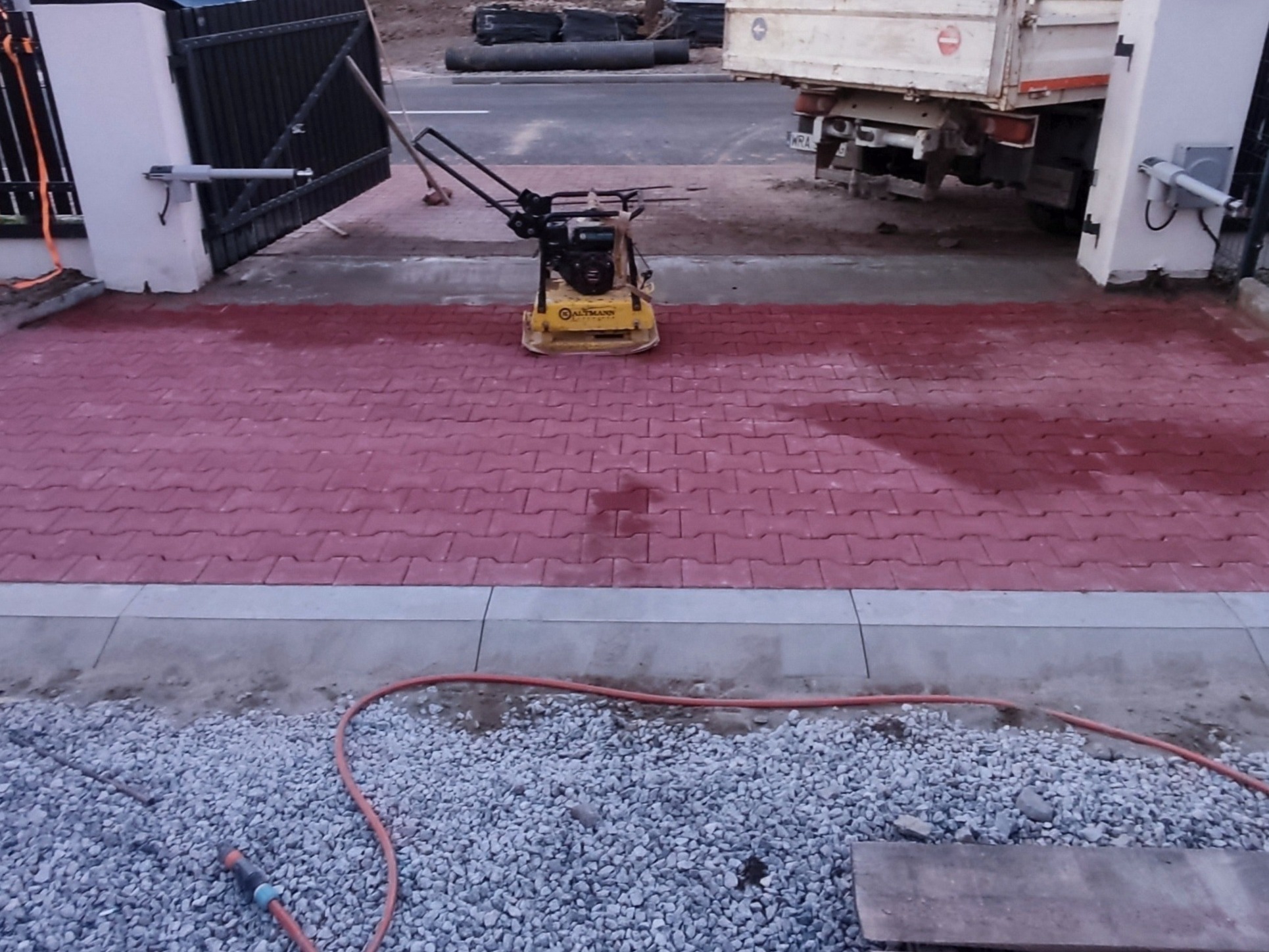 Czerwona kostka brukowa ułożona przy bramie, częściowo mokra po zagęszczaniu. W tle widoczna zagęszczarka i ciężarówka. Obrzeże oddziela kostkę od żwiru.