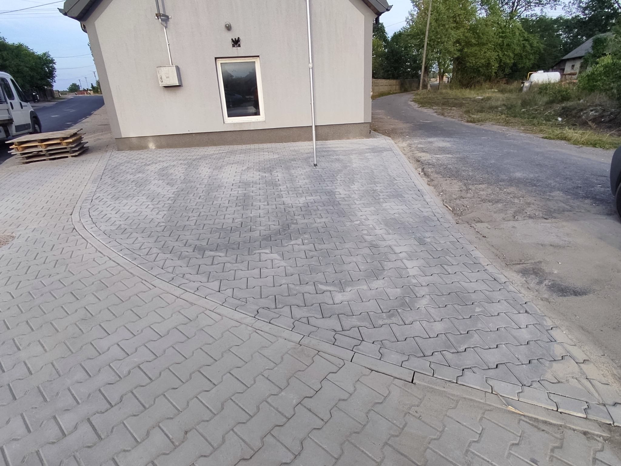 Szara kostka brukowa ułożona przed budynkiem z widocznymi paletami i fragmentem ulicy. Krawężnik oddziela chodnik od jezdni.
