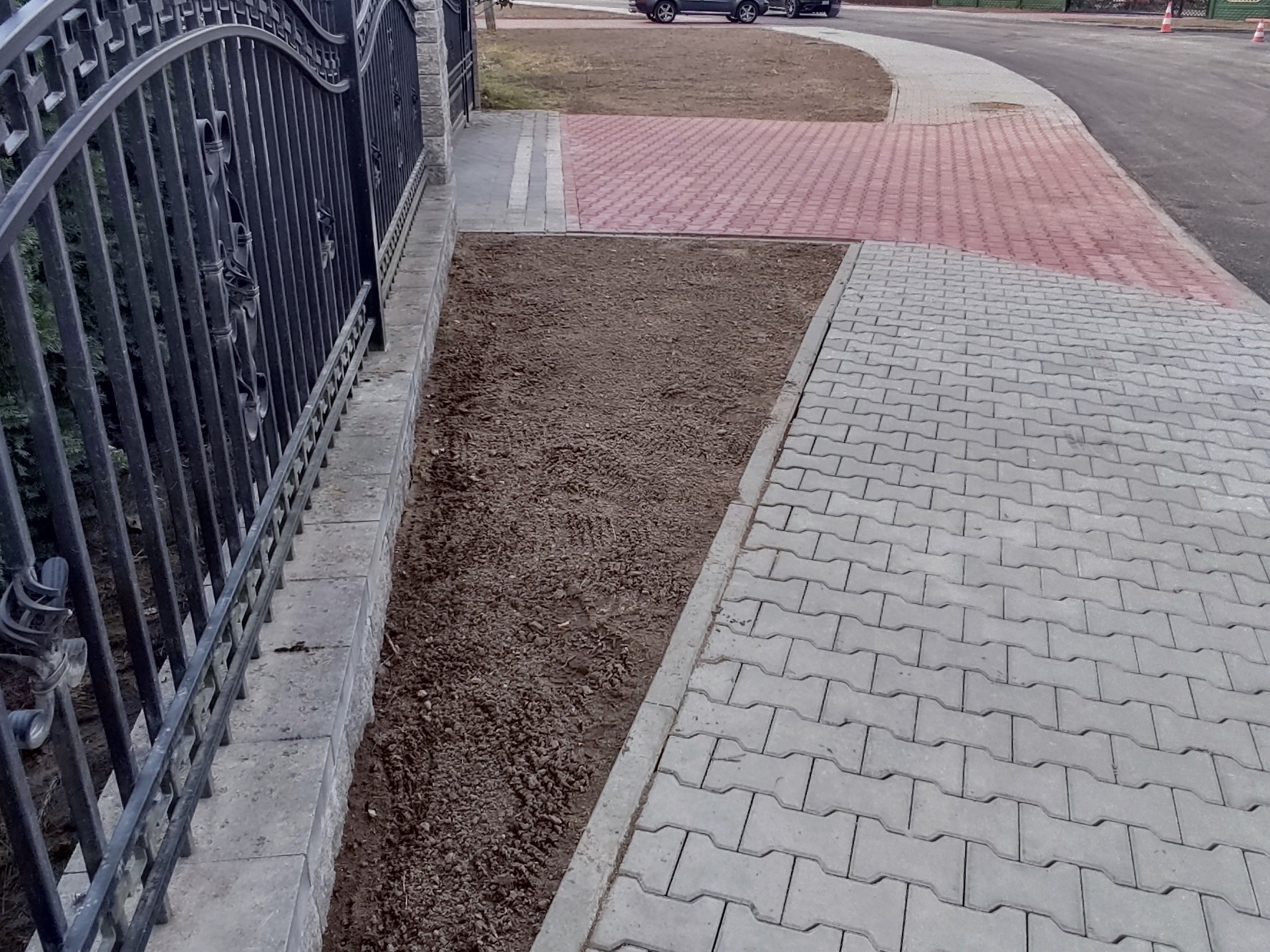 Ułożona szara kostka brukowa sąsiaduje z przygotowanym podłożem pod trawnik wzdłuż ozdobnego, kutego ogrodzenia. W tle widoczny fragment drogi z czerwonej kostki.