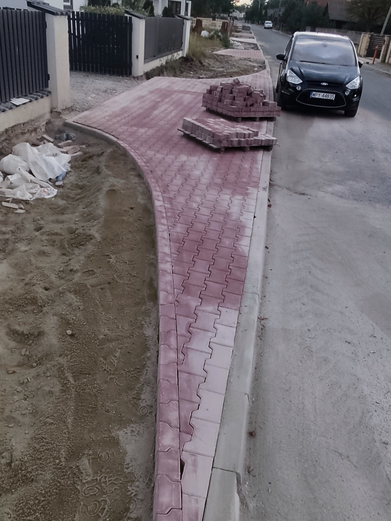 Fragment układanej, czerwonej kostki brukowej na chodniku z zaokrąglonym brzegiem, obok sterta kostki i czarny samochód. Widoczne przygotowane podłoże.