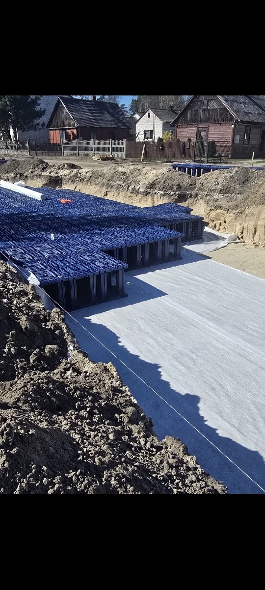 Przygotowany wykop z niebieską konstrukcją szalunkową i geowłókniną, gotowy do wylania fundamentów. W tle zabudowa wiejska w słoneczny dzień.