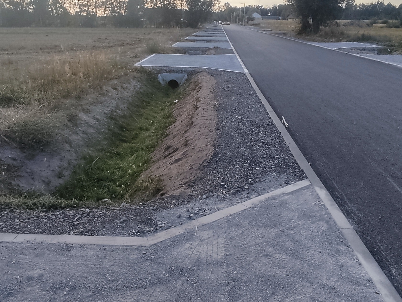 Nowa asfaltowa droga z wybrukowanym poboczem i rowem melioracyjnym z betonowymi wlotami. Widok na wprost, w tle zabudowania i drzewa.