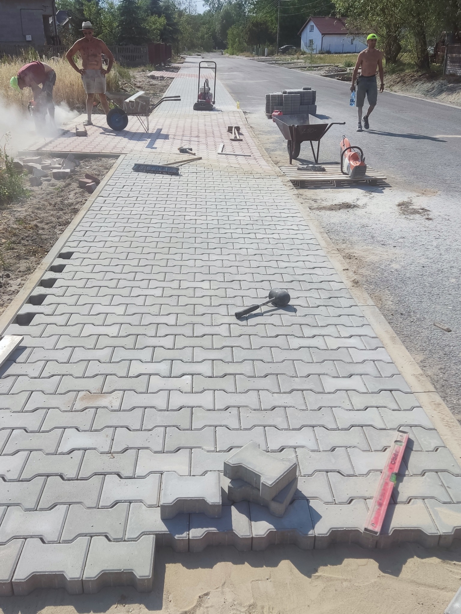 Układanie szarej kostki brukowej na chodniku w Szydłowcu. W tle pracownicy i narzędzia: taczka, zagęszczarka, piła do cięcia kostki. Widoczny postęp prac.