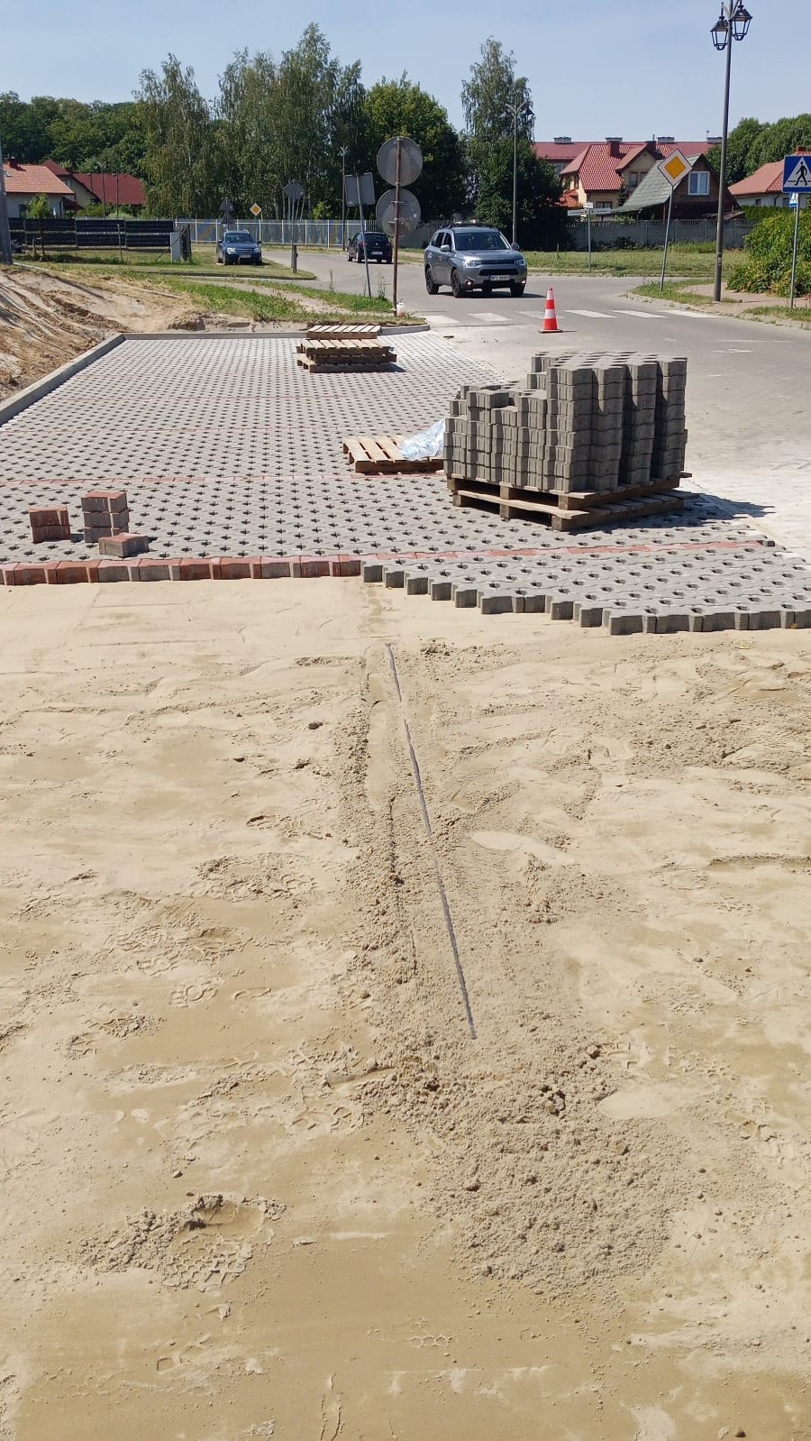 Fragment układanej nawierzchni z ażurowej kostki brukowej, paleta z kostką, przygotowane podłoże z piasku. W tle droga i zabudowa.