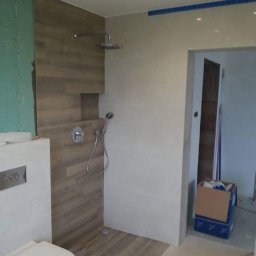 Krzysztof Rychlewski - Nowoczesna łazienka w trakcie remontu: kabina prysznicowa z drewnopodobnymi płytkami, podtynkowa bateria, toaleta i surowa ściana z zieloną płytą gipsową.