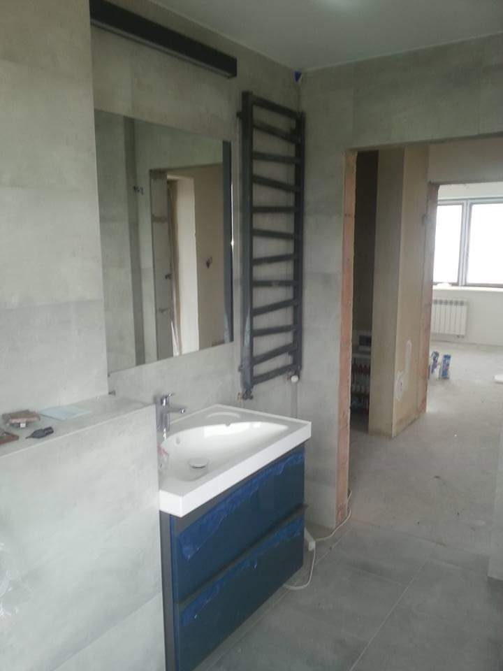 Łazienka w trakcie wykończenia: umywalka z szafką, grzejnik drabinkowy, płytki na ścianach i podłodze. Widoczne elementy zabezpieczające przed zabrudzeniem.