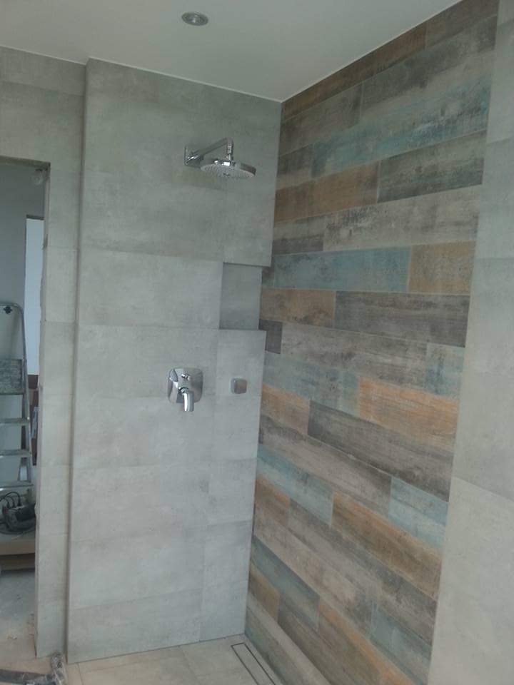 Nowoczesna kabina prysznicowa z szarymi płytkami imitującymi beton i ścianą akcentową z płytek drewnopodobnych w odcieniach brązu i błękitu. Widoczna armatura i odpływ liniowy.