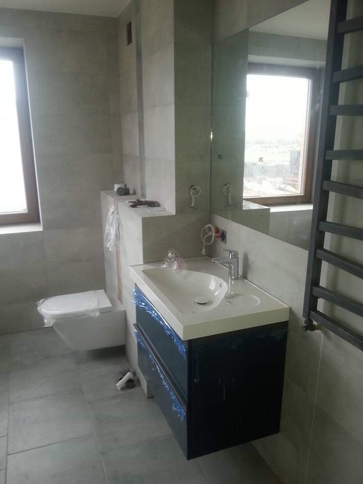 Nowoczesna łazienka w szarościach: podwieszana toaleta, szafka z umywalką i lustro. Elementy zabezpieczone folią ochronną. Minimalistyczny design, płytki imitujące kamień.