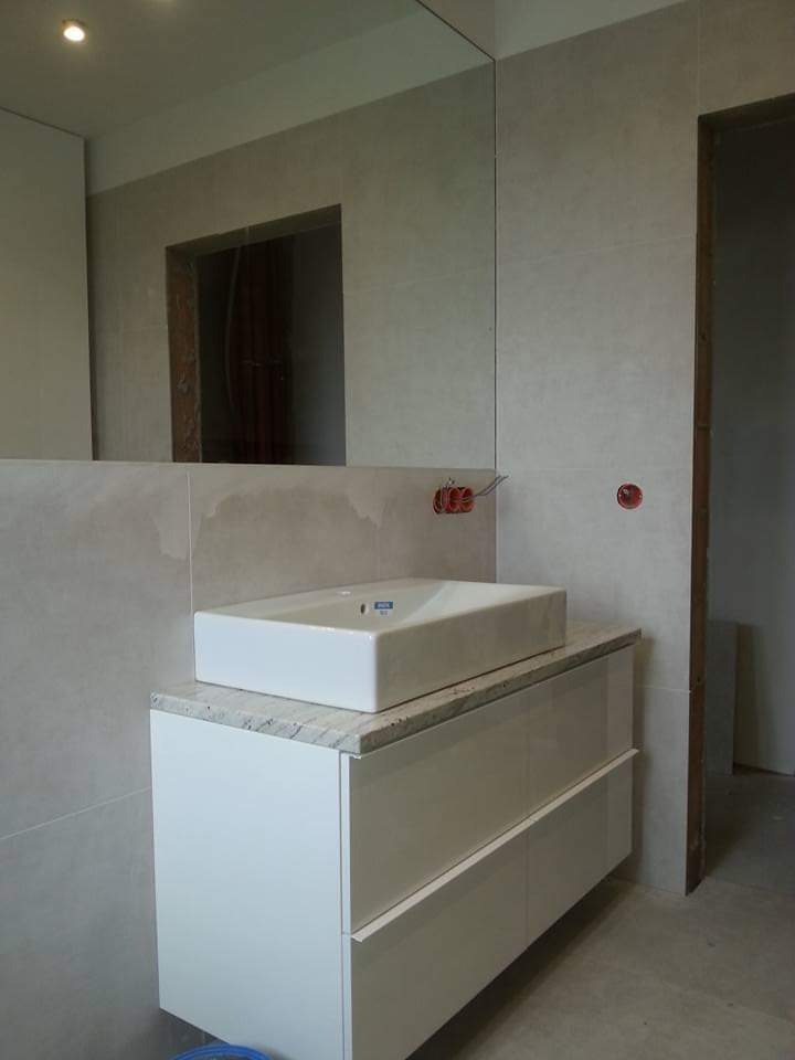 Nowoczesna łazienka z umywalką nablatową na białej szafce z blatem imitującym kamień. W tle lustro na całą ścianę i szare płytki. Widoczne gniazdka elektryczne.