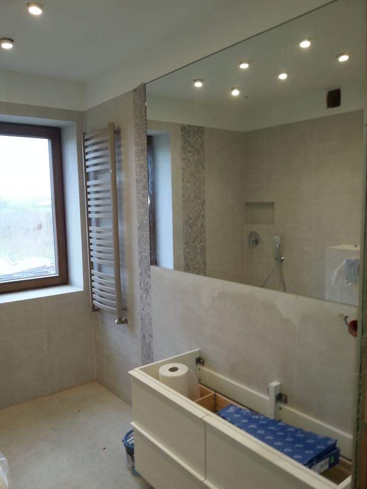 Jasna łazienka z beżowymi płytkami, dużym lustrem i grzejnikiem drabinkowym. Umywalka w trakcie montażu, widoczne akcesoria. Oświetlenie sufitowe punktowe.