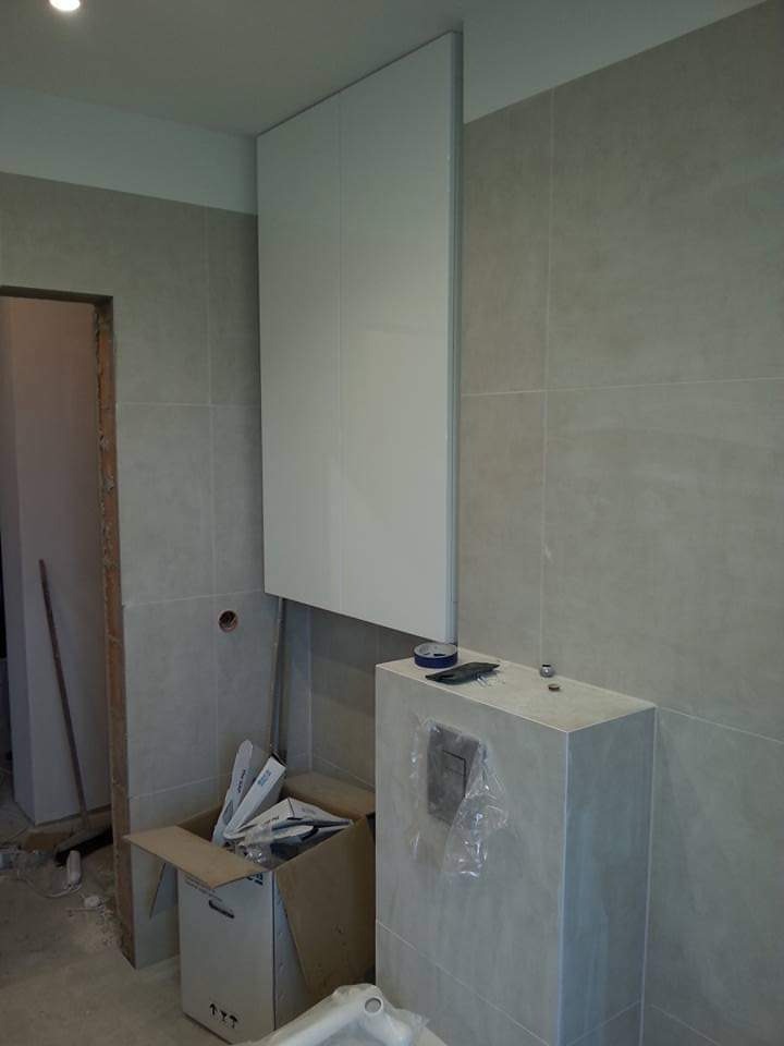 Ściana w łazience wyłożona płytkami w odcieniach szarości. Widoczna zabudowa stelaża WC i szafka wisząca. Na podłodze karton z akcesoriami. Etap prac wykończeniowych.