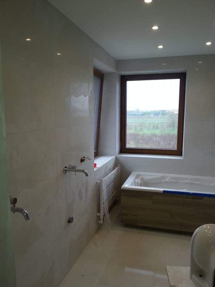 Jasna łazienka z oknem: beżowe płytki, drewniana obudowa wanny, chromowana armatura. Widok na zewnątrz przez okno. Grzejnik drabinkowy w folii ochronnej.