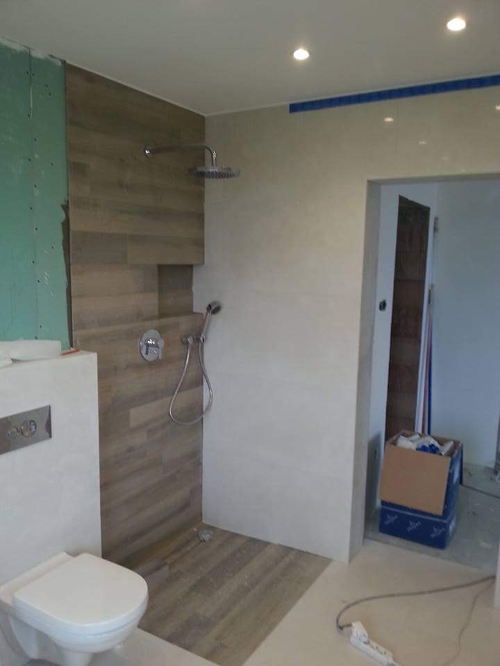 Nowoczesna łazienka w trakcie remontu: kabina prysznicowa z drewnopodobnymi płytkami, podtynkowa bateria, toaleta i surowa ściana z zieloną płytą gipsową.