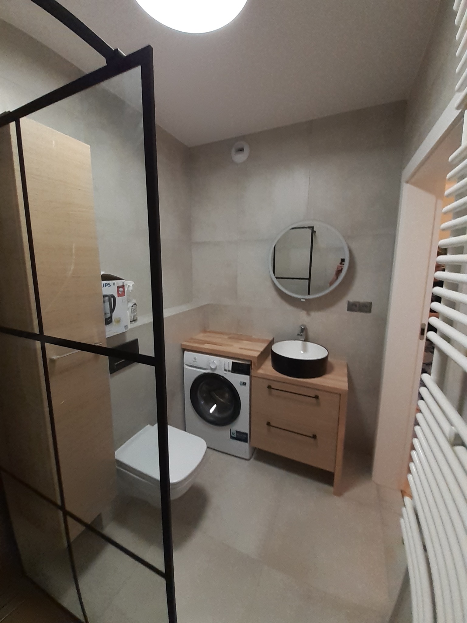 Jasna łazienka z kabiną prysznicową typu walk-in, umywalką na szafce z drewnianym blatem, pralką i okrągłym lustrem. Minimalistyczny design, neutralna kolorystyka.