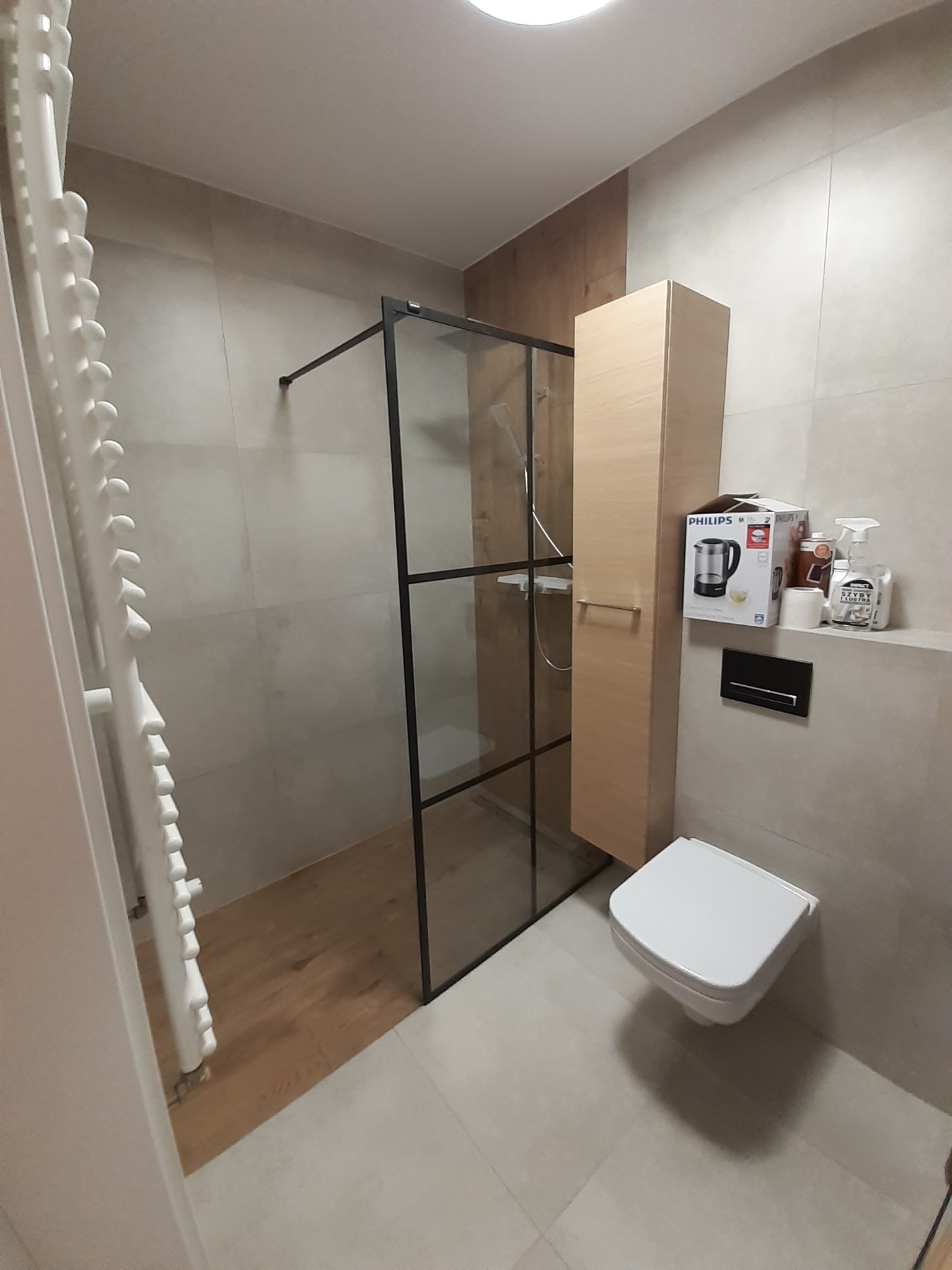 Nowoczesna łazienka z kabiną prysznicową typu walk-in z czarnymi profilami, wiszącą toaletą i grzejnikiem drabinkowym. Beżowe płytki i drewniane akcenty.