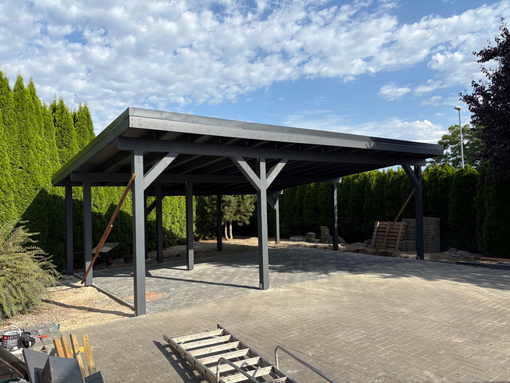 Nowoczesna, grafitowa pergola z drewnianą konstrukcją na tle zielonych iglaków i błękitnego nieba. Widoczne narzędzia sugerują ukończenie montażu.