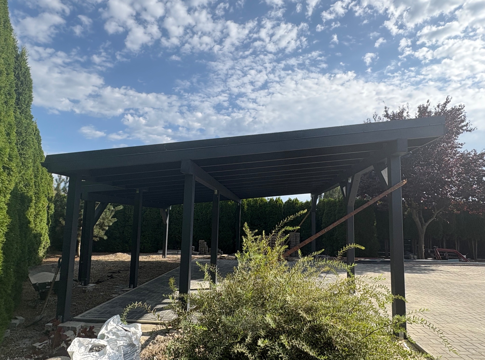 Nowoczesna, czarna pergola z drewnianą konstrukcją na tle błękitnego nieba z chmurami. Widoczne elementy ogrodu i kostka brukowa.