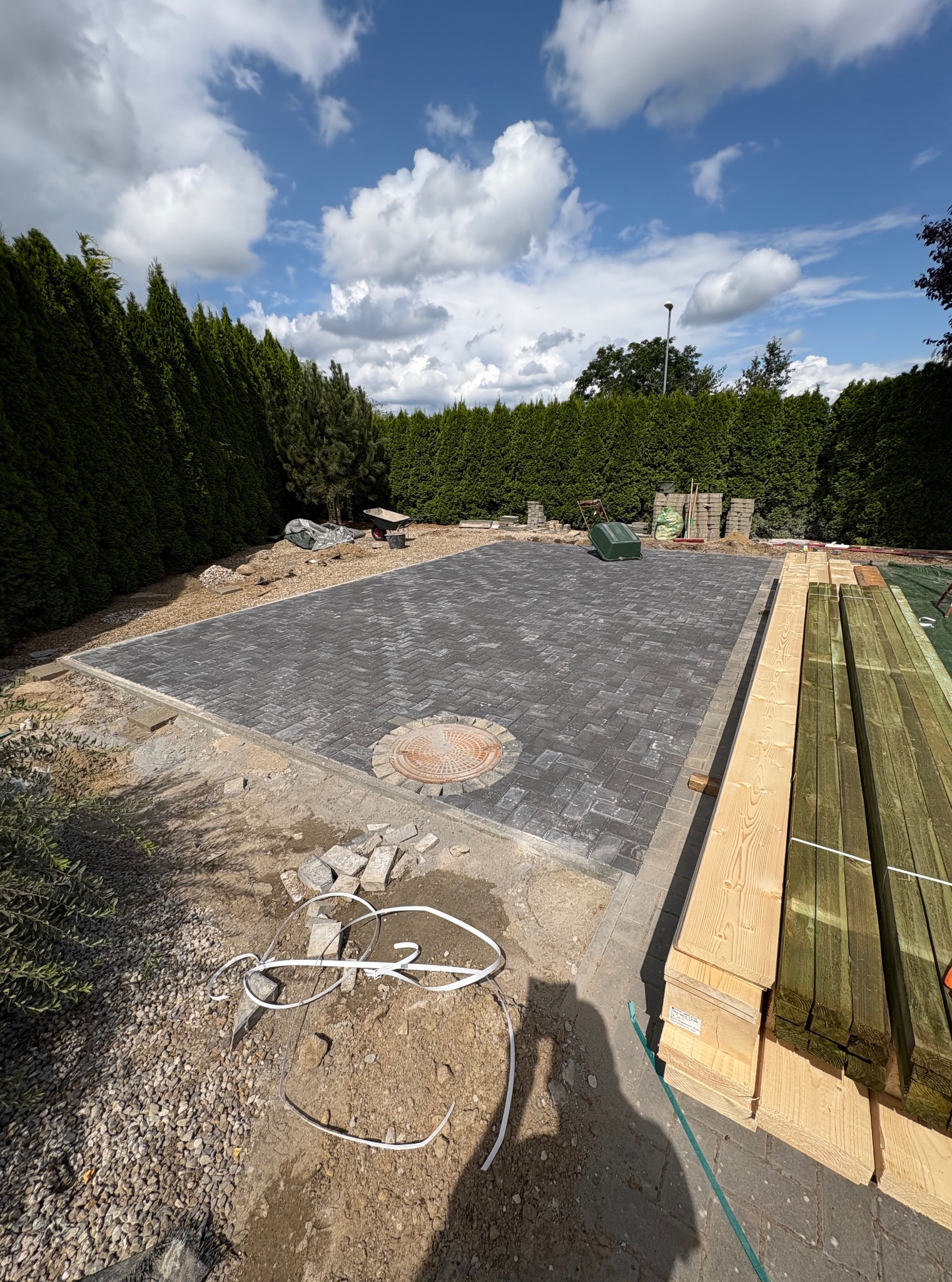 Układanie kostki brukowej na tarasie w ogrodzie, widoczne przygotowane drewniane deski. W tle zielone tuje i błękitne niebo z chmurami. Prace w toku.