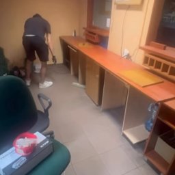 Usługi Utylizacji Busem Jakub Molenda - Sprzątanie biura: mężczyzna zamiata podłogę wzdłuż długiego, drewnianego biurka. Widoczne krzesło biurowe i akcesoria. Ujęcie z perspektywy podłogi, lekko z dołu.