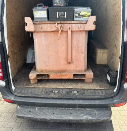 Metalowy kontener na palecie i akumulatory w przestrzeni ładunkowej białego busa. Widok z tyłu pojazdu, gotowy do transportu.