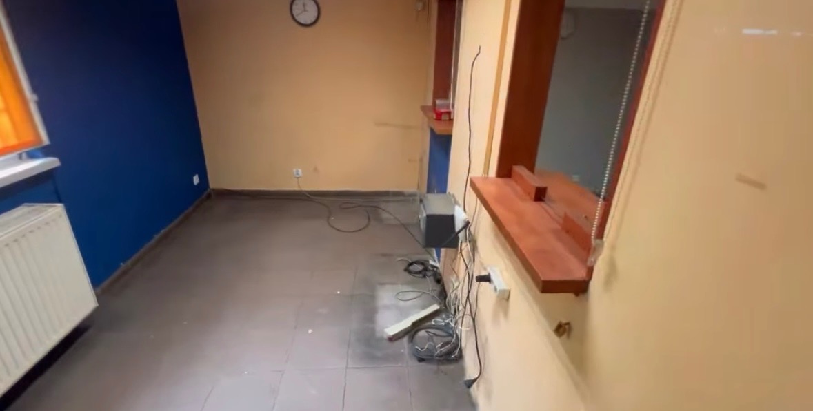 Pusta przestrzeń biurowa z niebieską ścianą, brązową podłogą i widocznymi kablami na podłodze. Na ścianie wisi zegar. Widoczne okienko podawcze.