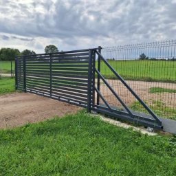 P.H.U HURTOWNIA STALI JUSTYNA KOBUS - Nowoczesna, przesuwna brama wjazdowa z metalowymi, poziomymi panelami, widoczna na tle zielonego trawnika i siatki ogrodzeniowej. Brama w kolorze grafitowym.