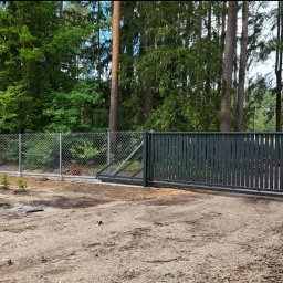 P.H.U HURTOWNIA STALI JUSTYNA KOBUS - Kompletne ogrodzenie posesji z siatki i przesuwną bramą w kolorze grafitowym, na tle leśnego krajobrazu. Widoczne elementy montażowe na ziemi.