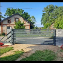 Nowoczesna, szara brama wjazdowa z poziomymi panelami, zamontowana na tle domu z cegły i zieleni. Widoczne elementy automatyki i solidne słupki.