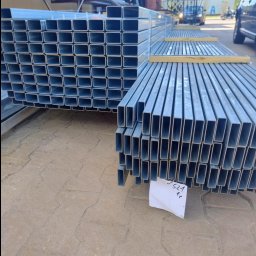 P.H.U HURTOWNIA STALI JUSTYNA KOBUS - Stosy metalowych profili o przekroju prostokątnym, ułożonych na zewnątrz. Widoczne elementy konstrukcyjne budynku i pojazdy w tle. Na pierwszym planie kartka z numerem.