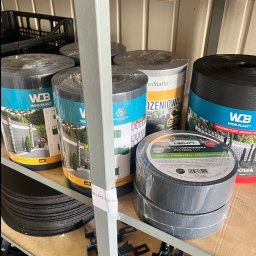 P.H.U HURTOWNIA STALI JUSTYNA KOBUS - Rolki taśmy ogrodzeniowej PVC Heun antracyt i panele WDB Plast z grafiką ogrodzeń na półce magazynowej. Elementy montażowe i kratki w tle.