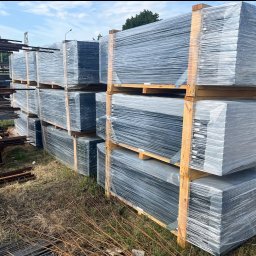 P.H.U HURTOWNIA STALI JUSTYNA KOBUS - Stos paneli ogrodzeniowych, zabezpieczonych folią i ułożonych na drewnianych paletach. W tle widać składowane metalowe elementy konstrukcyjne.