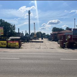 P.H.U HURTOWNIA STALI JUSTYNA KOBUS - Widok z ulicy na plac firmy oferującej ogrodzenia, bramy i akcesoria. W tle widoczny dźwig i wysoki komin. Banery reklamowe przy wjeździe.