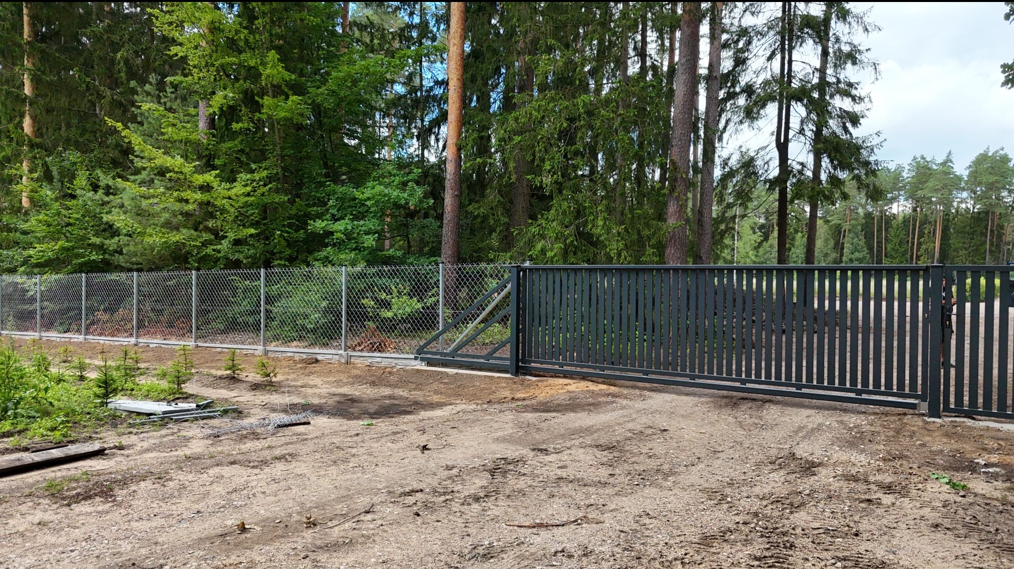 Kompletne ogrodzenie posesji z siatki i przesuwną bramą w kolorze grafitowym, na tle leśnego krajobrazu. Widoczne elementy montażowe na ziemi.