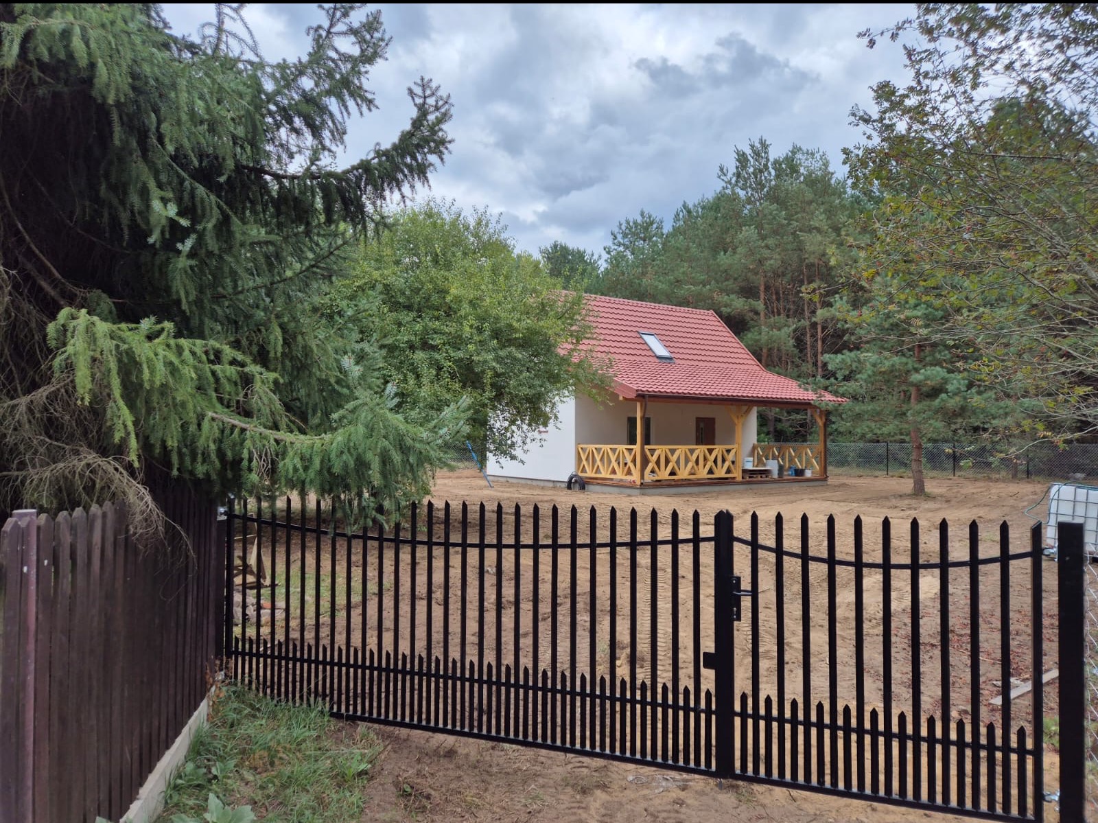 Czarny metalowy płot sztachetowy przed domem z czerwoną dachówką, widok zza świerku. Na posesji piasek, w tle las.