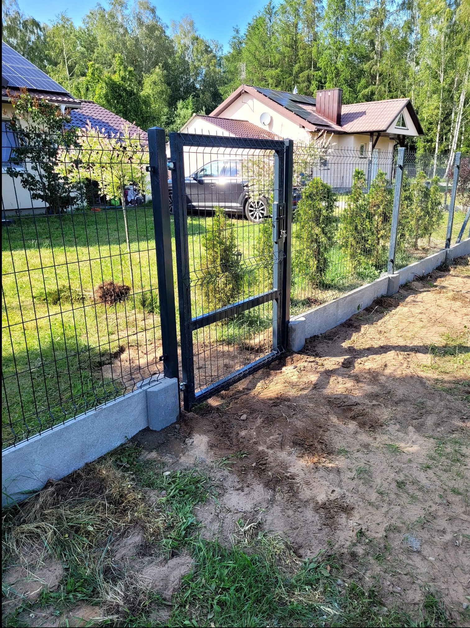 Nowoczesne ogrodzenie panelowe z furtką, osadzone na betonowej podmurówce, w tle dom z panelami solarnymi i zielony ogród. Słoneczny dzień.