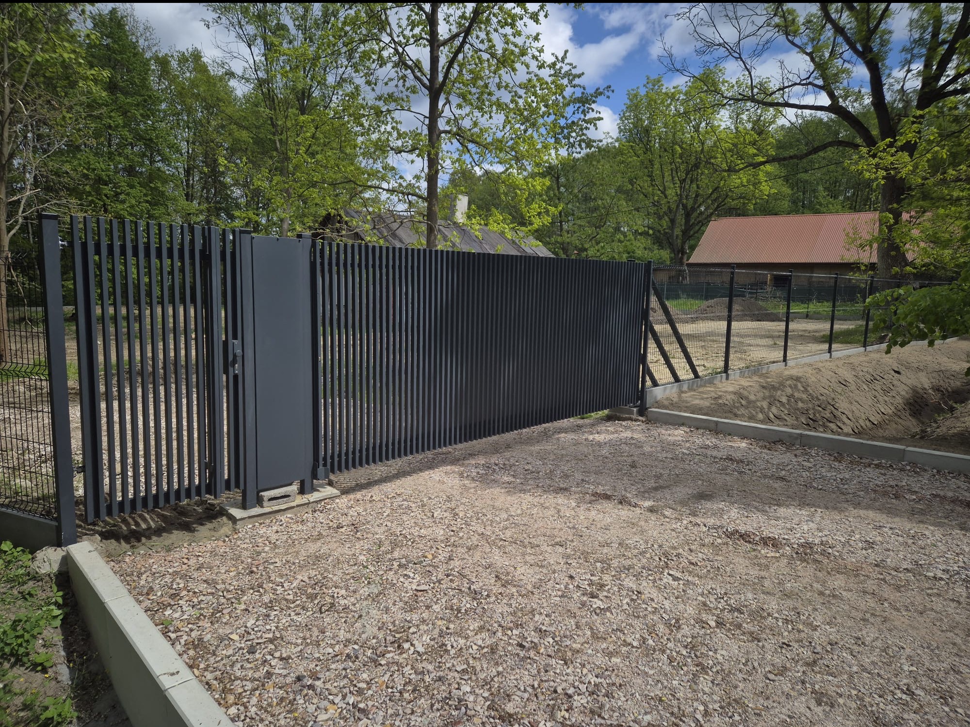 Nowoczesna, metalowa brama przesuwna w kolorze grafitowym, z pionowymi listwami, na tle zieleni drzew i żwirowego podjazdu. Widoczny fragment ogrodzenia z siatki.