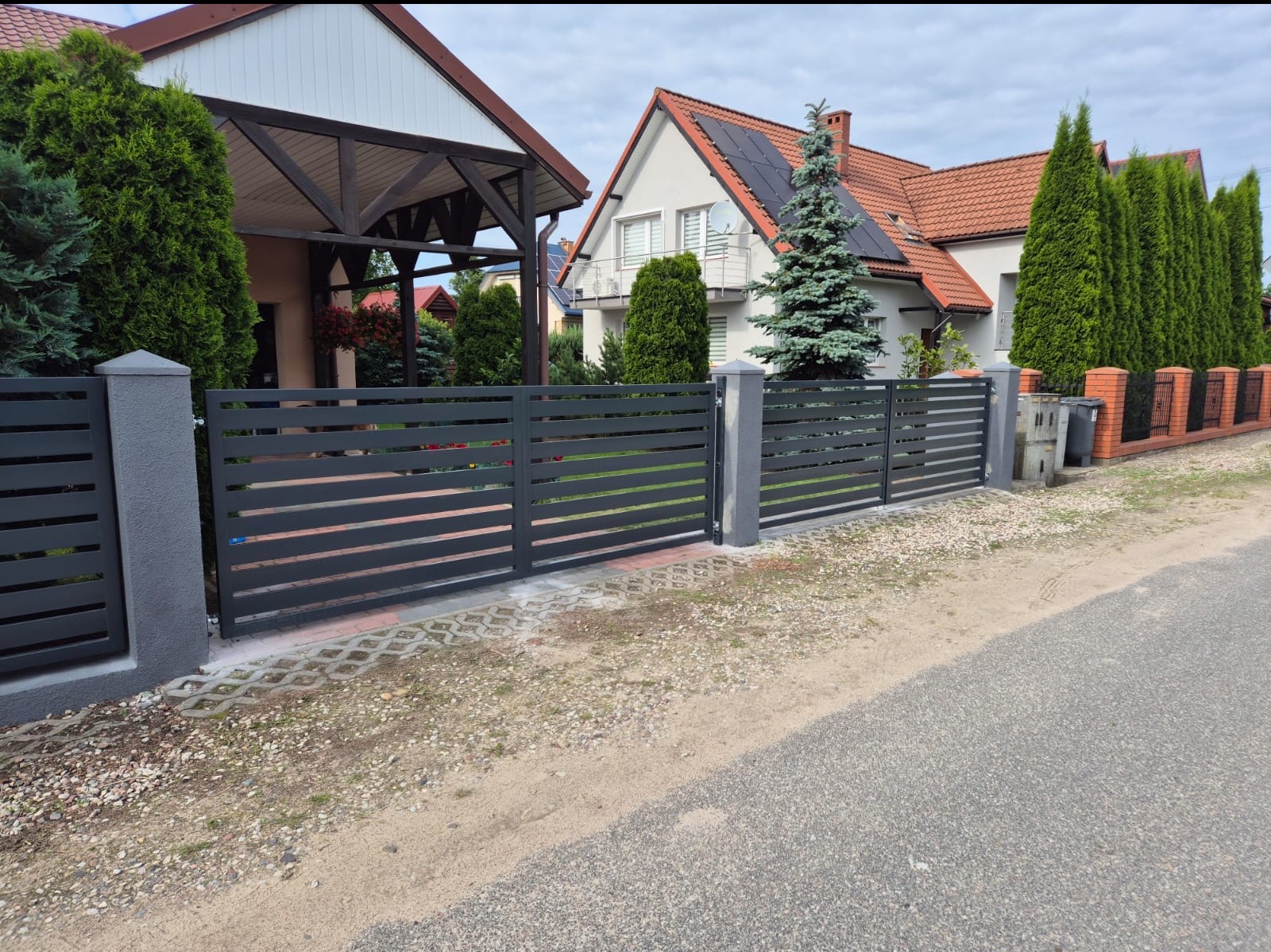 Nowoczesne, poziome przęsła z szarego metalu tworzą ogrodzenie posesji z kostki ażurowej. W tle dom z panelami słonecznymi na dachu.
