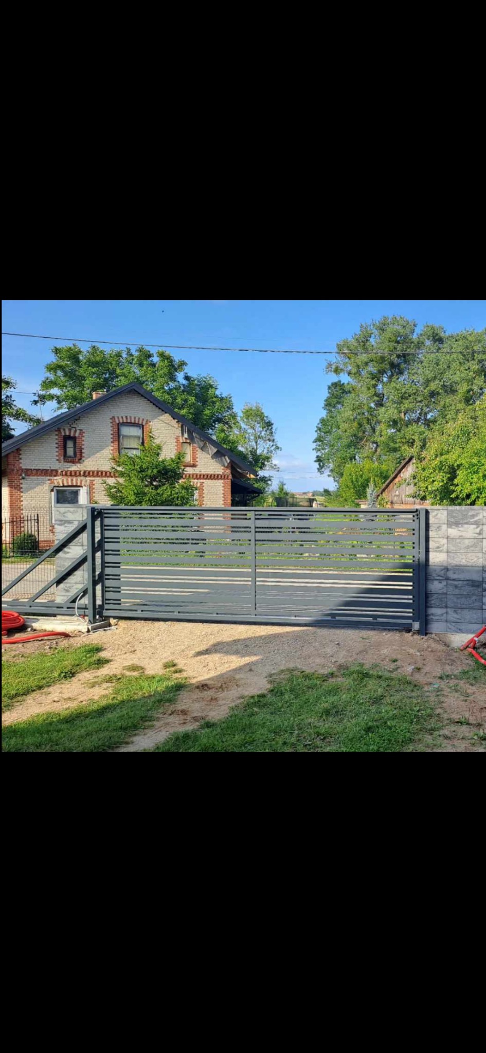 Nowoczesna, szara brama wjazdowa z poziomymi panelami, zamontowana na tle domu z cegły i zieleni. Widoczne elementy automatyki i solidne słupki.