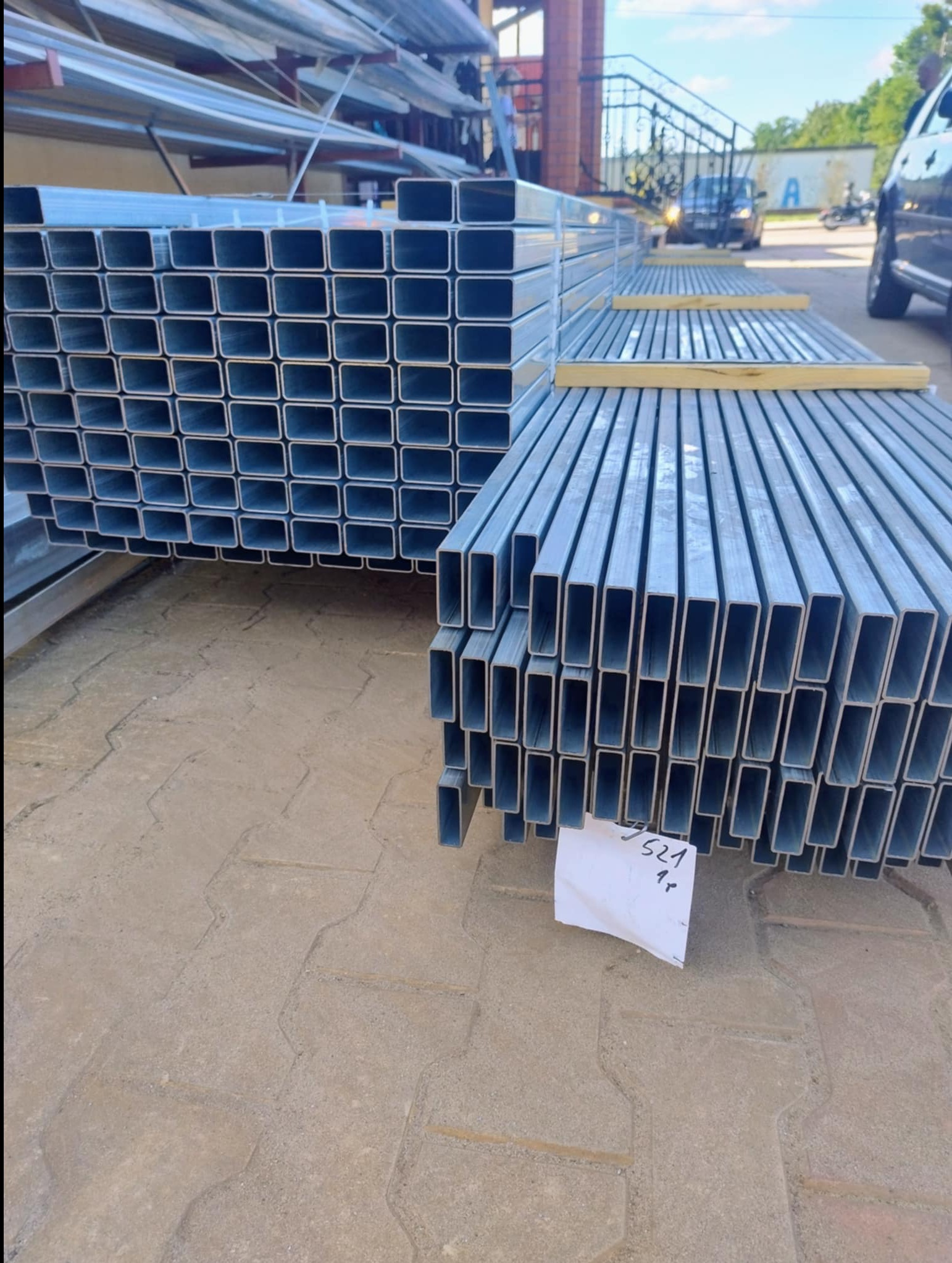 Stosy metalowych profili o przekroju prostokątnym, ułożonych na zewnątrz. Widoczne elementy konstrukcyjne budynku i pojazdy w tle. Na pierwszym planie kartka z numerem.