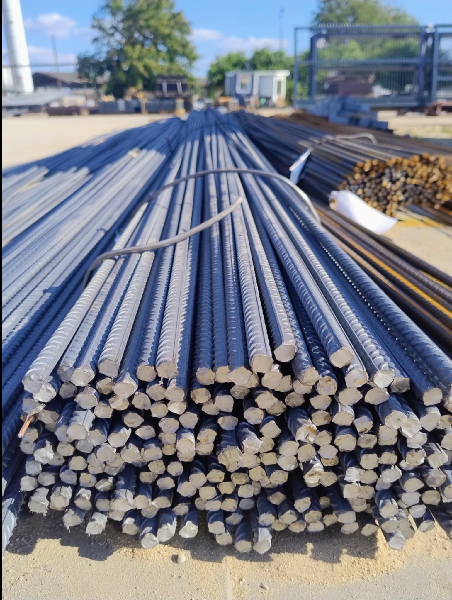 Stalowe pręty zbrojeniowe ułożone w stos, widoczne poprzeczne przekroje. W tle brama wjazdowa i budynek gospodarczy. Materiał budowlany na placu.