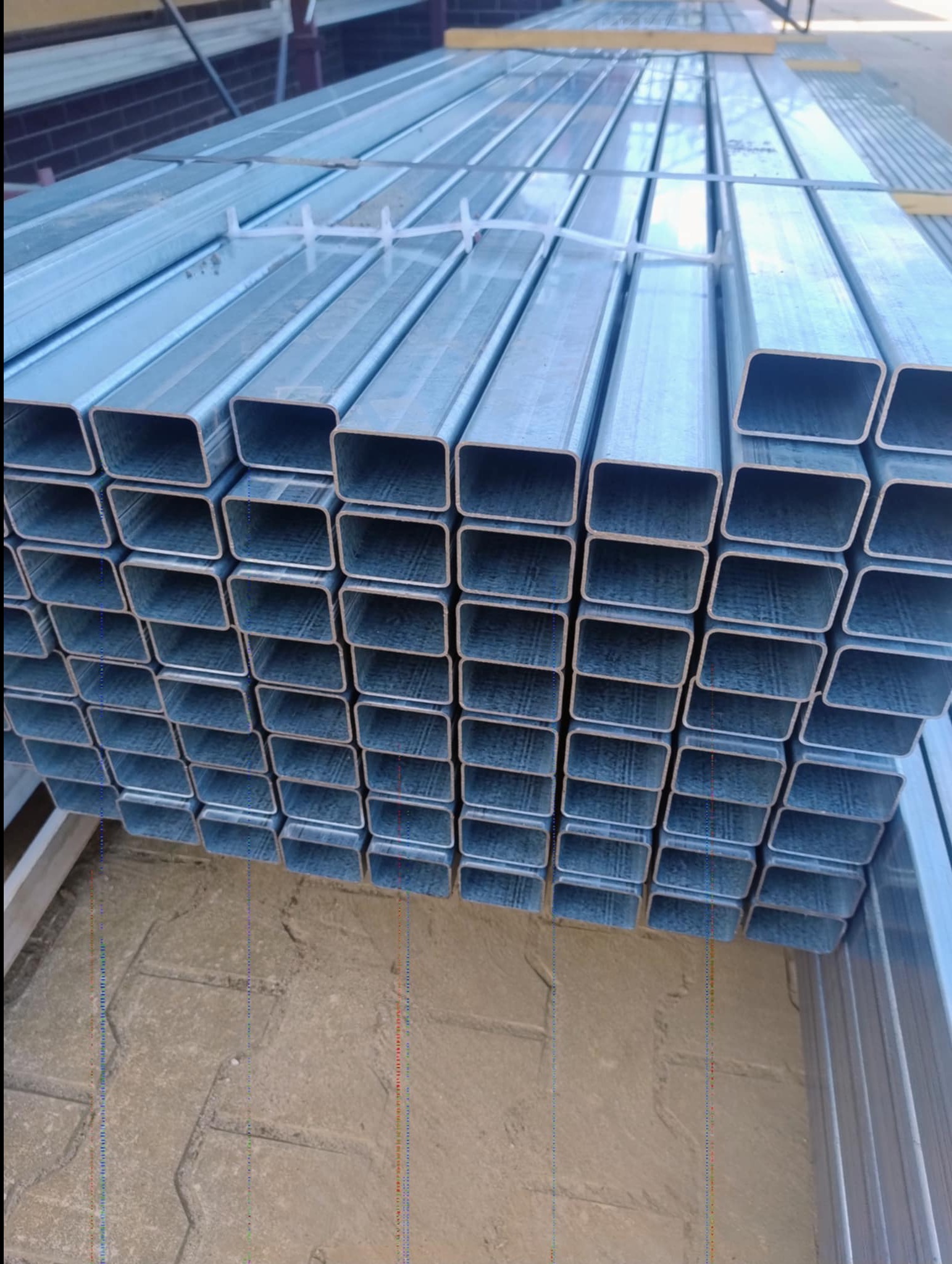 Stalowe, prostokątne profile ogrodzeniowe ułożone w stos. Elementy zabezpieczone plastikowymi taśmami. Widoczny fragment kostki brukowej w tle.