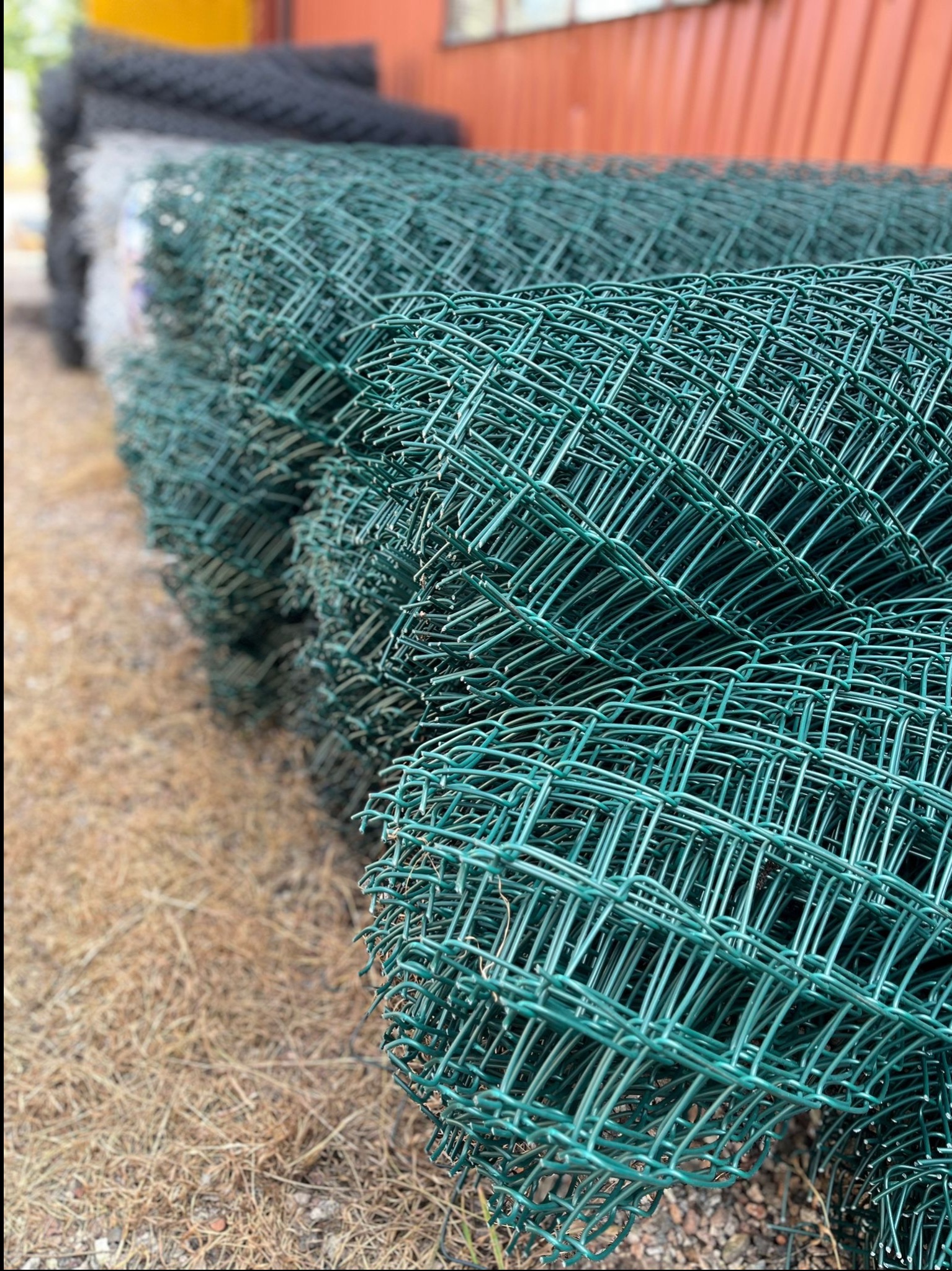 Zrolowana, zielona siatka ogrodzeniowa na ziemi, z widocznymi oczkami i fakturą drutu. W tle, inne rolki siatki i fragment budynku.