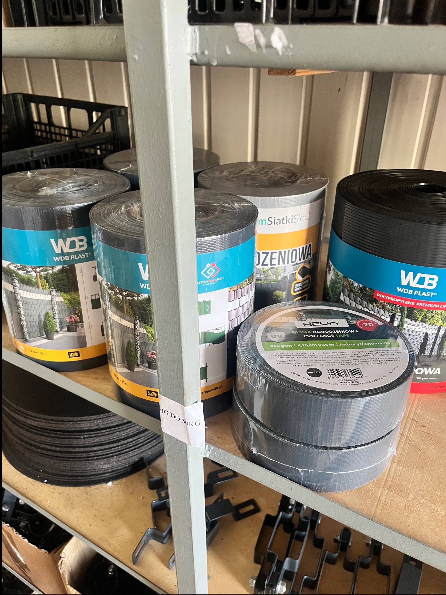 Rolki taśmy ogrodzeniowej PVC Heun antracyt i panele WDB Plast z grafiką ogrodzeń na półce magazynowej. Elementy montażowe i kratki w tle.