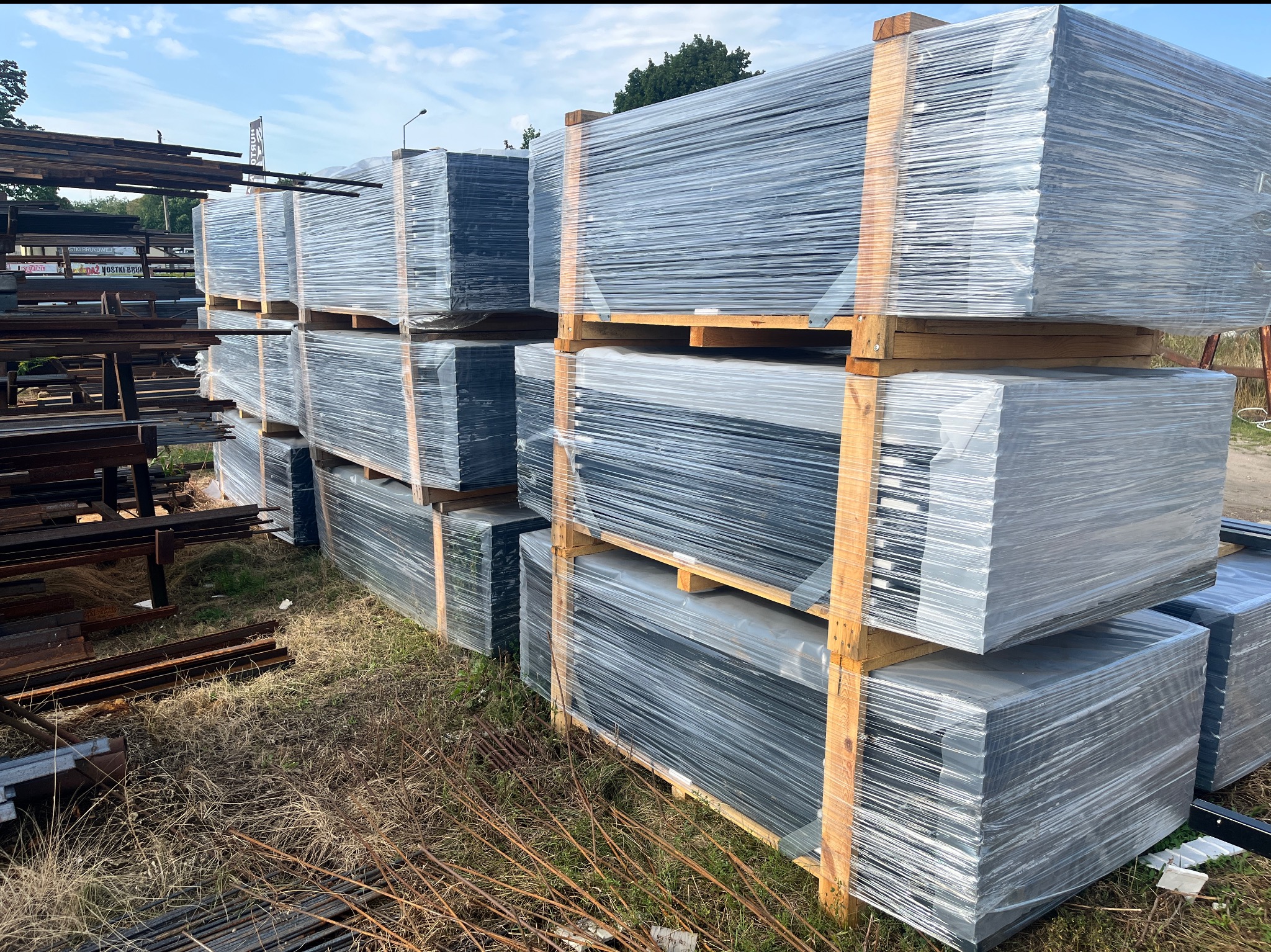 Stos paneli ogrodzeniowych, zabezpieczonych folią i ułożonych na drewnianych paletach. W tle widać składowane metalowe elementy konstrukcyjne.