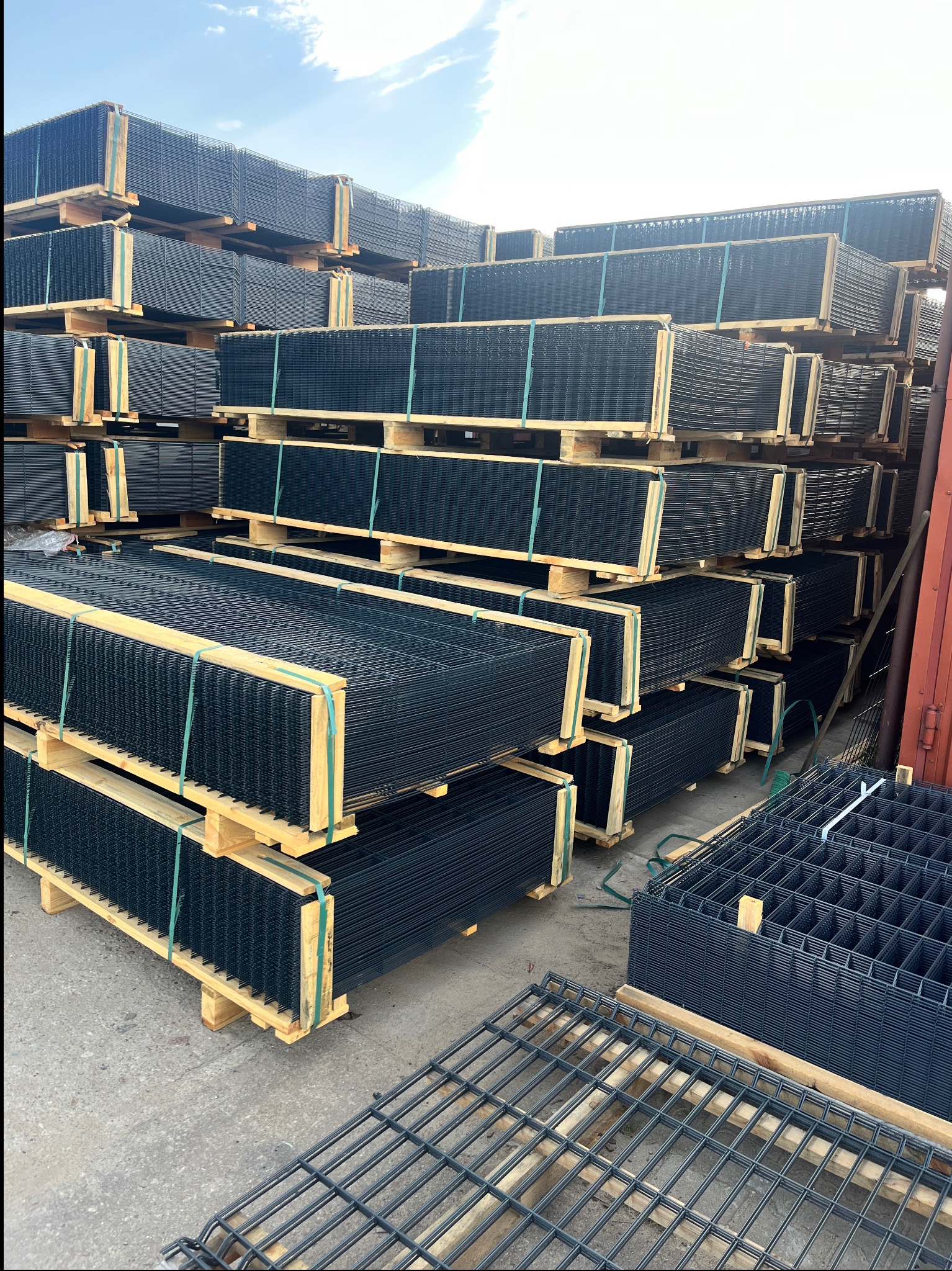 Stos palet z panelami ogrodzeniowymi w kolorze grafitowym, zabezpieczonych taśmami. Na pierwszym planie fragment panelu. Widoczne niebo i fragment ściany.