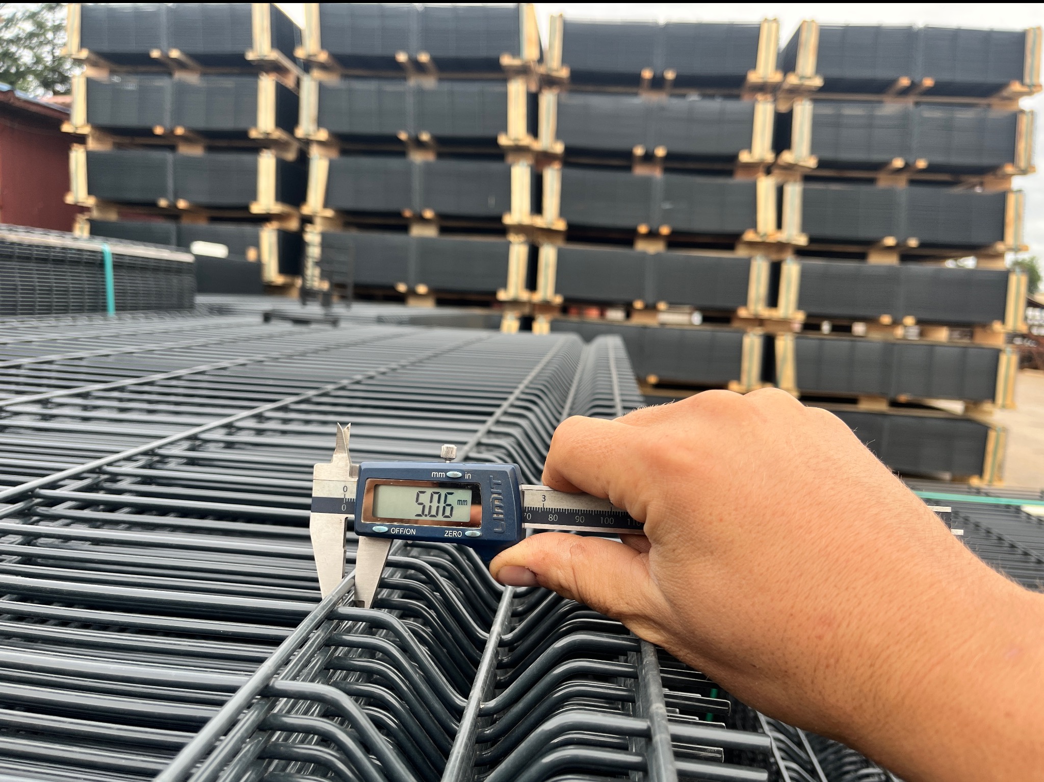 Pomiar grubości drutu panelu ogrodzeniowego suwmiarką elektroniczną, stos paneli w tle. Szary panel zgrzewany, widoczny wynik 5.06 mm.