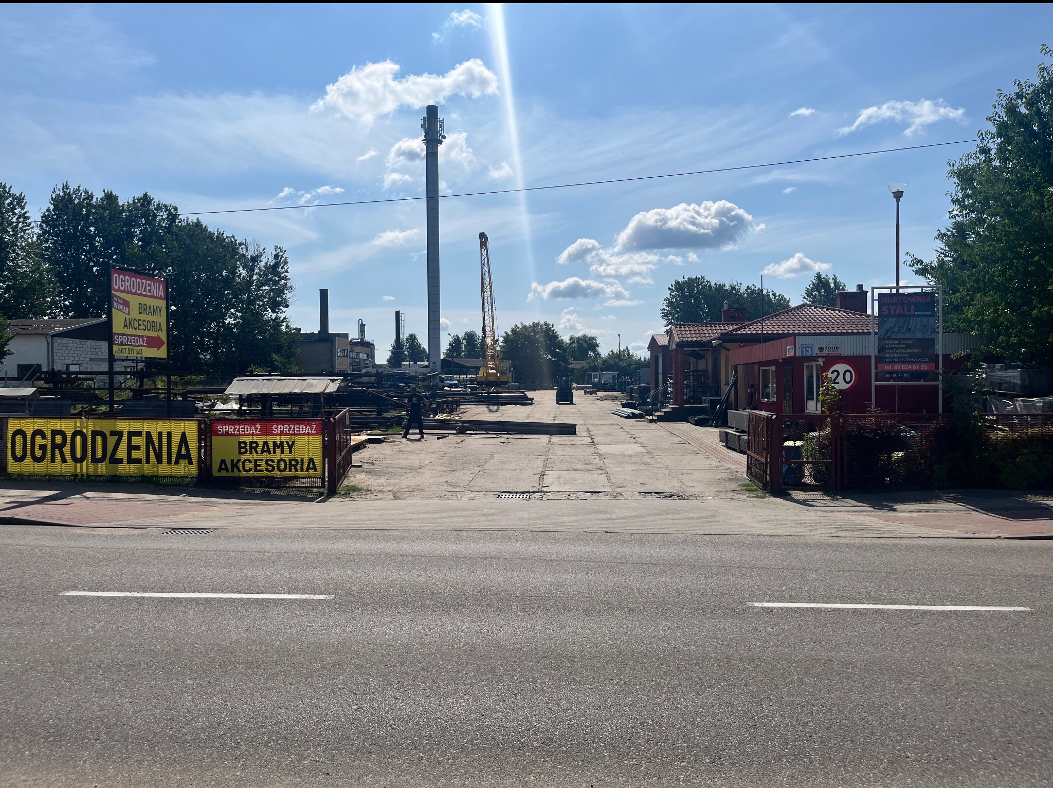 Widok z ulicy na plac firmy oferującej ogrodzenia, bramy i akcesoria. W tle widoczny dźwig i wysoki komin. Banery reklamowe przy wjeździe.