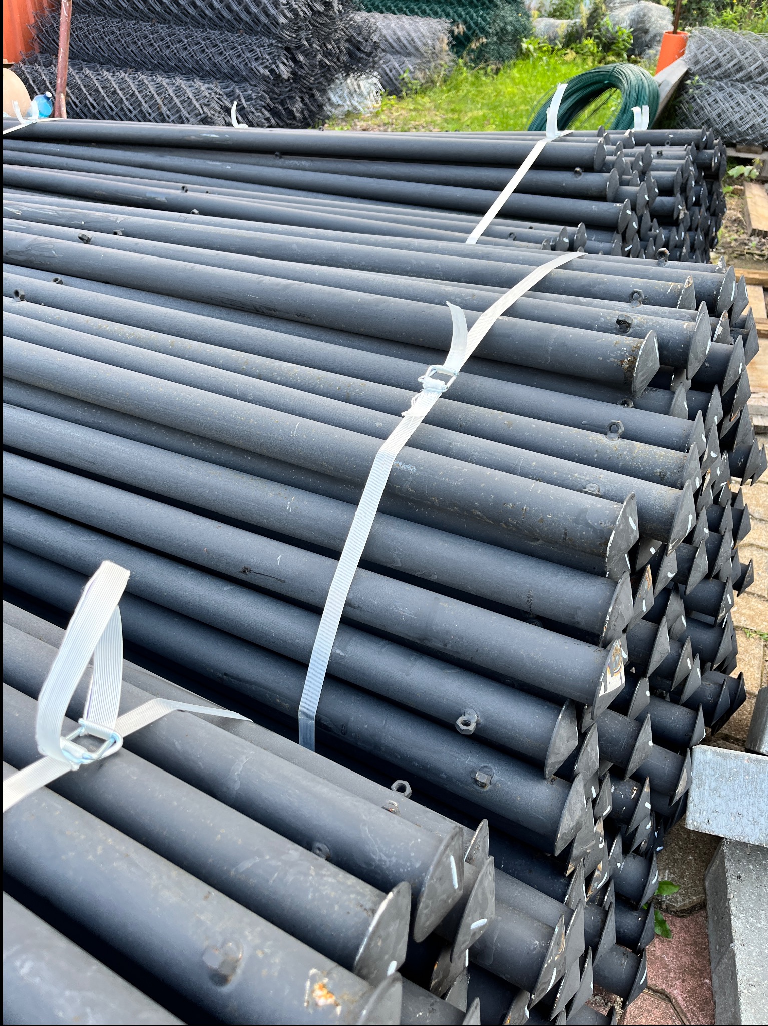 Stos słupków do montażu ogrodzeń panelowych w Szczytnie, spiętych taśmą. Widoczne kotwy i oczka montażowe. W tle siatka ogrodzeniowa i trawa.